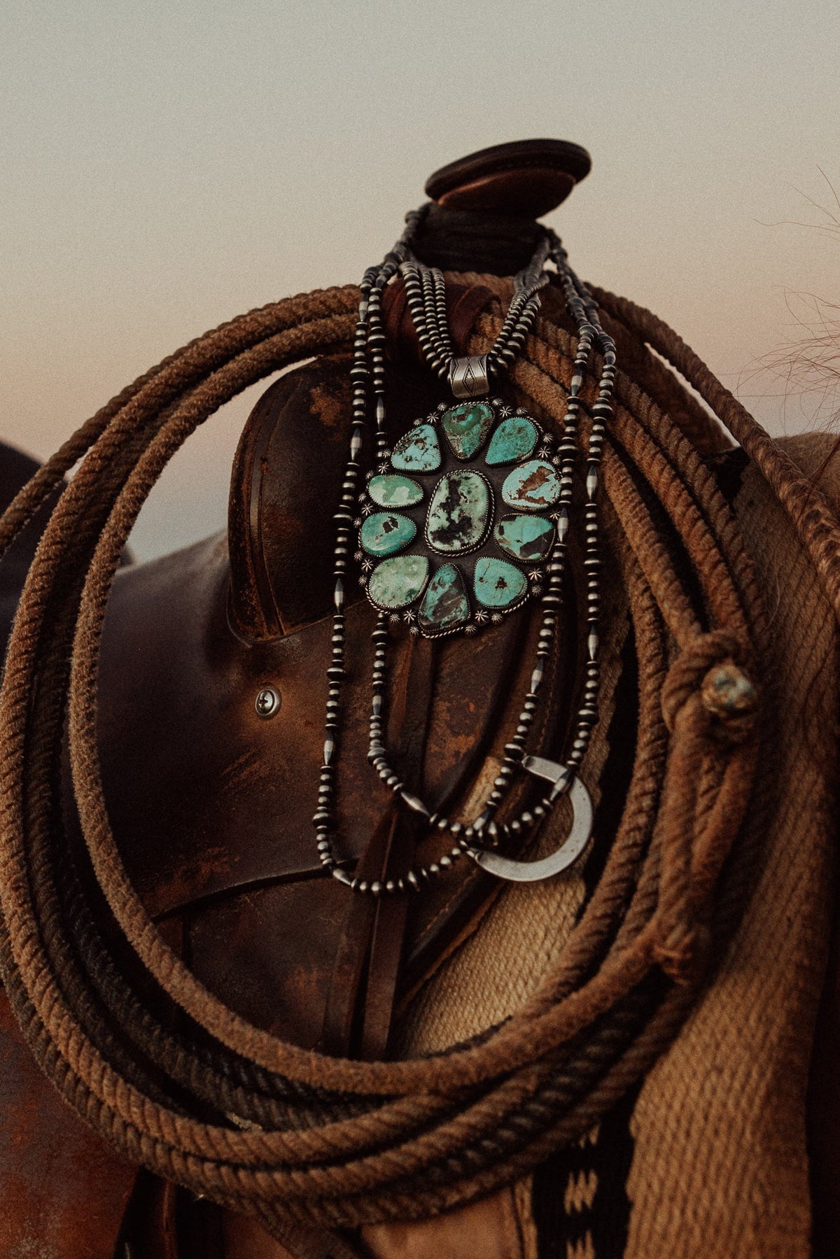 A close-up of a brown leather saddle decorated with multiple strands of silver beadwork and a large turquoise stone cluster pendant, with coiled brown rope draped around it against a warm beige sky.