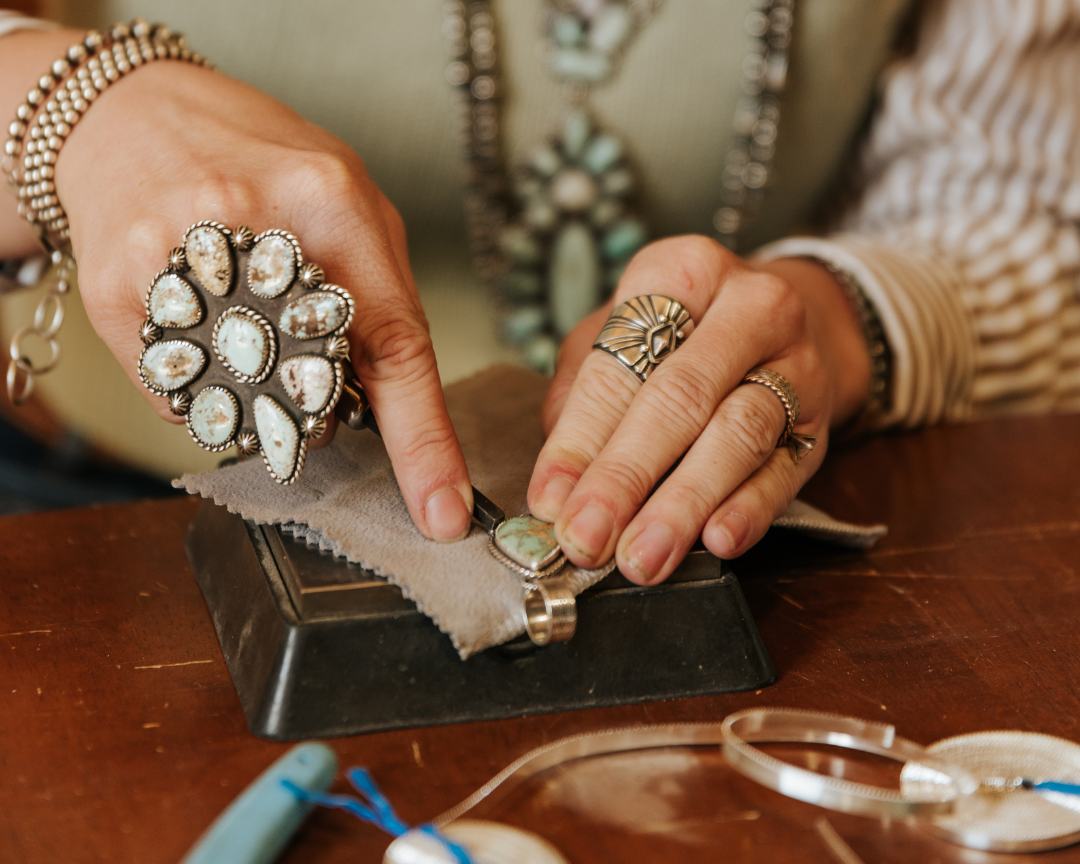 A close-up of a person's hands crafting jewelry on a wooden workbench, wearing multiple silver-toned rings and a silver-toned necklace with turquoise-colored stones.
