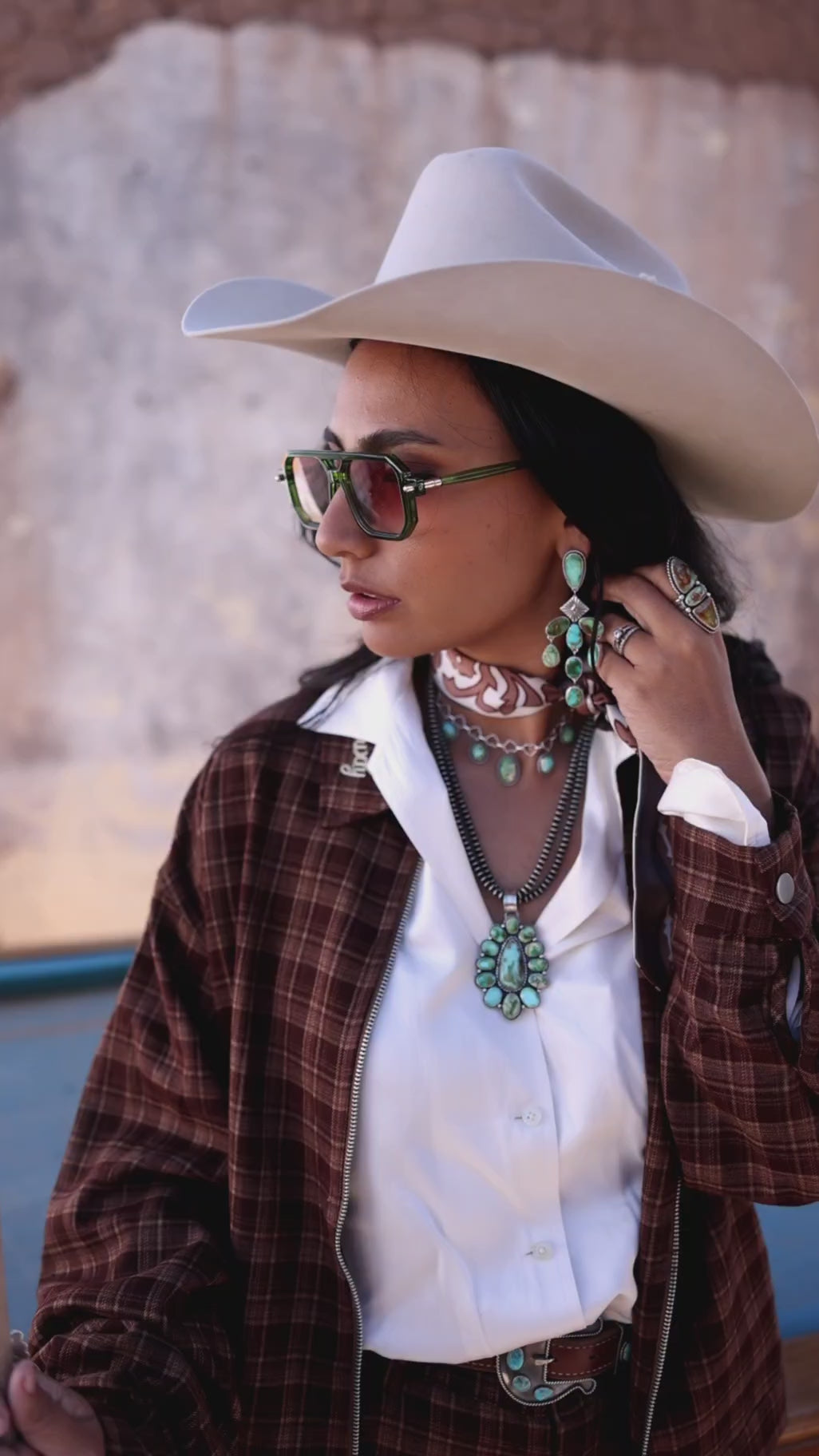  A cowgirl models handmade turquoise jewelry from Double M Beadwork, styled with a white cowboy hat, sunglasses, white shirt, and brown plaid suit. Close-up shots highlight the turquoise earrings and statement pendant, followed by wider views of the complete Western look. Handmade in West Texas for cowgirls and women in the Western industry. 