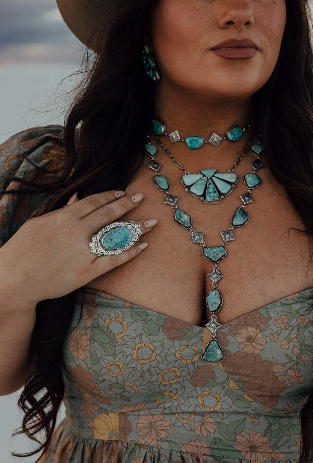 A person wearing a floral dress and a wide-brim hat is adorned with a cluster-style necklace featuring white water turquoise stones, along with matching earrings and a ring.